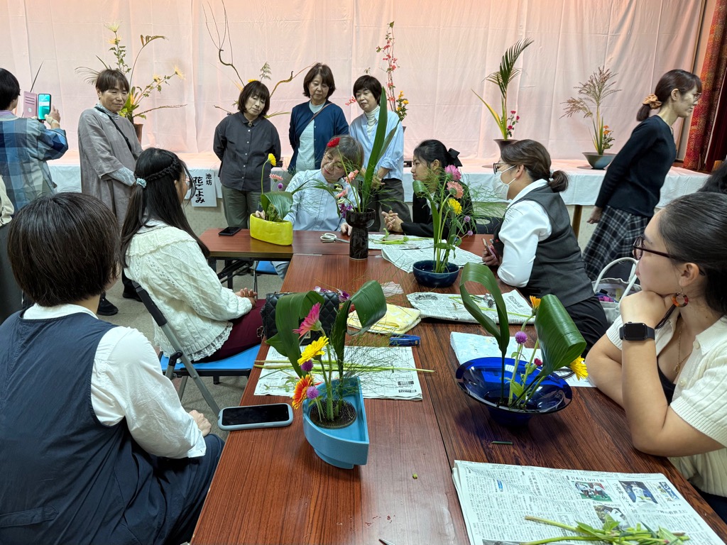 生けた花を先生に見てもらう女性を周囲の女性たちが見守っている